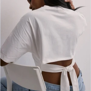 Vit croppad topp med knyt i ryggen - Säljer en vit croppad topp med korta ärmar och snygg knytning i ryggen. Toppen har en enkel och clean look framtill, men baksidan har en öppen detalj med breda band som knyts till en rosett. Perfekt till jeans för en chill och trendig stil.