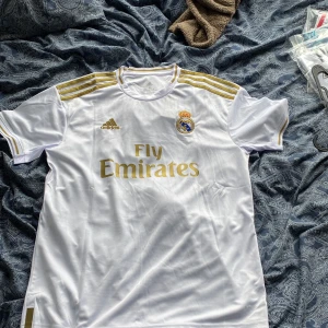 Real Madrid Benzema tröja XL Adidas - Snygg Real Madrid matchtröja från Adidas i vitt med guldiga detaljer och Benzema 9 på ryggen. Tröjan har korta ärmar, broderat klubbmärke och är tillverkad i lätt och ventilerande Climalite-material. Perfekt för dig som vill visa ditt stöd för Los Blancos. Tröjan är helt i nyskick och lappar sitter även kvar. Storleken är XL så passar någon som är 180-185 cm lång. Vid minsta fråga eller om du vill ha mer bilder på tröjan hör av dig.