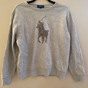 Grå sweatshirt från Polo Ralph Lauren - Snygg grå sweatshirt från Polo Ralph Lauren med det klassiska stora polospelar-loggan framtill. Tröjan har rund hals, långa ärmar och ribbade muddar vid ärmslut och nederkant. Perfekt för en chill och stilren look.
