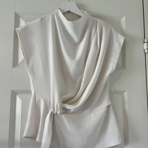 Vit blus från Zara - Stilren vit blus med lös passform. Toppen har korta ärmar och en snygg drapering framtill som ger en cool och modern look. Perfekt för dig som gillar minimalistisk stil och vill ha något unikt i