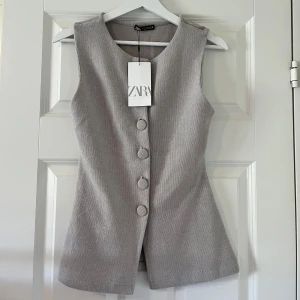 Grå västopp från Zara - Snygg grå väst från Zara med figurnära passform och stora klädda knappar framtill. Västen är ärmlös och har en stilren, minimalistisk design som ger en clean look. Perfekt att styla över en skjorta eller t-shirt för en trendig vibe.