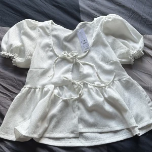 Vit blus med puffärm och knyt - Superfin vit blus Gina med puffiga korta ärmar och två knyt framtill. Blusen har en lätt textur i tyget och volang nedtill helt oanvänd