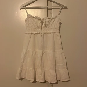 Vit kort sommarklänning från Bershka - Supersöt vit kort klänning från Bershka i storlek XS. Klänningen har smala axelband, volangdetaljer upptill och vid fållen samt knytning framtill. Tillverkad i ett lätt och luftigt material, perfekt för varma dagar.