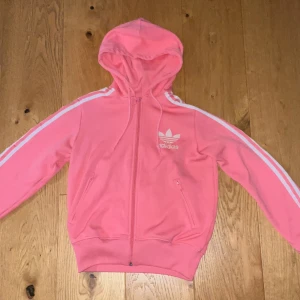Retro adidas Hoddie  - Säljer en rosa Adidas hoodie med vit logga och klassiska ränder längs ärmarna. Jackan har huva, hel dragkedja och två fickor framtill. Perfekt för en sportig och chill stil. Materialet är mjukt och skönt!😃