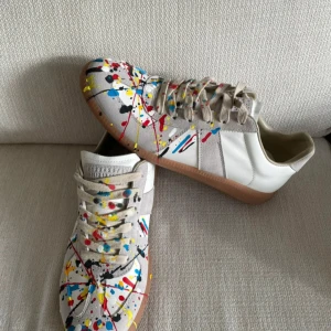 Maison Margiela sneakers med färgstänk - Unika sneakers från Maison Margiela i vitt och beige skinn och mocka, dekorerade med färgstänk i rött, blått, gult och svart. Klassisk låg modell med snörning och gummisula. Perfekt för dig som vill sticka ut med en kreativ vibe.