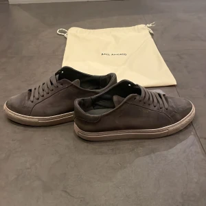 Gråa sneakers från Axel Arigato - Snygga gråa sneakers från Axel Arigato i mocka med klassisk låg profil och rund tå. Skorna har matchande gråa snören och en beige sula med diskret logga på sidan. Perfekta för dig som gillar stilrena och minimalistiska skor.