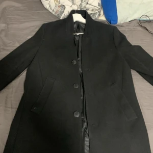 Svart klassisk kappa med knappar - Stilren svart kappa med rak passform och diskret krage. Den har en liten slits på baksidan. Använt knappast