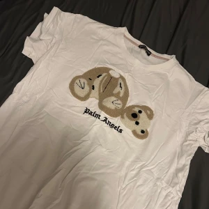 Vit Palm Angels t-shirt med nalle - Vit t-shirt från Palm Angels med ett stort broderat motiv av en nallebjörn på bröstet och logotyp under. Klassisk rund hals och korta ärmar. Perfekt för dig som vill sticka ut med en unik och trendig design.
