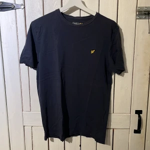 Mörkblå t-shirt från Lyle & Scott - Snygg mörkblå t-shirt från Lyle & Scott med klassisk gul logga på bröstet. Enkel och stilren design med rund hals och korta ärmar. Perfekt för dig som gillar en clean look och vill ha ett plagg som funkar till det mesta. Är en L men sitter som en M
