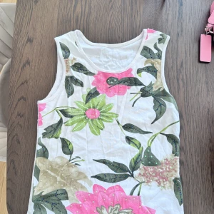 Blommig ärmlös linnetopp med paljetter - Superfin ärmlös linnetopp med stora blommor i rosa, grönt och beige på vit botten. Toppen har glittriga paljettdetaljer som ger extra wow-känsla. Perfekt för dig som gillar färg och vill sticka ut lite extra i sommar.