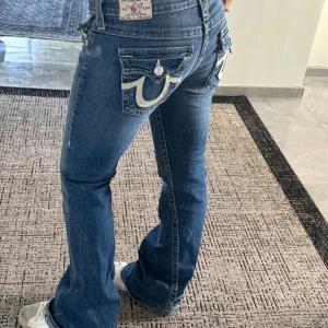 True Religion blå bootcut jeans - Säljer ett par blå jeans från True Religion med ikoniska vita sömmar och stora fickor bak. Modellen har låg midja och bootcut-ben, klassisk tvättad denim och snygga detaljer på fickorna. Perfekta för dig som gillar street och retrovibe.kolla mått för storlek!!!! Innerbenslängd: 84cm