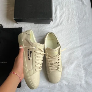 Saint Laurent Sneakers - Säljer ett par ÄKTA vita/beige sneakers från Saint Laurent med handskriven logga på sidan. Skorna har rund tå, platt sula och är tillverkade i canvas med vita snören. Klassisk och stilren design med diskreta detaljer. Storlek 41 sitter som 41. Skick 10/10. Tillbehör/OG Låda och 2 dustbags.
