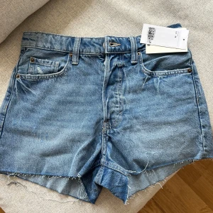 Blå jeansshorts från H&M - Snygga blå jeansshorts från H&M med råa, fransiga kanter och klassisk femficksdesign. Modellen är högmidjad och har en avslappnad passform. Perfekta för varma dagar och enkel att matcha med allt. Ikonisk denimlook som aldrig går ur tiden.