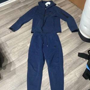 Mörkblå nike tech set - Snygga mörkblå mjukisbyxor från Nike med dragsko i midjan och diskret logga på benet. Sköna och stilrena, perfekta för chill eller träning. Tillverkade i mjukt material som känns bekvämt hela dagen.