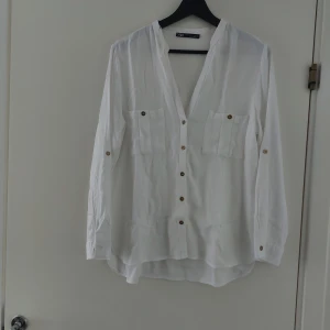 Vit v-ringad skjorta från Zara - Vit skjorta från Zara med v-ringning, långa ärmar och snygga bruna knappar framtill. Två bröstfickor och lätt, flowigt material som ger en chill vibe. Perfekt för dig som gillar stilrena plagg med enkel design.