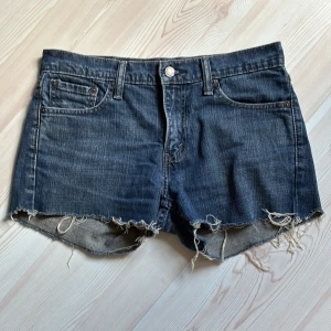 Levi's jeansshorts med fransig kant - Mörkblå jeansshorts från Levi's med klassisk femficksdesign och rå, fransig kant nertill. Shortsen har normal passform och är perfekta för sommaren. Snygg slitning och ikonisk Levi's-detalj bak.