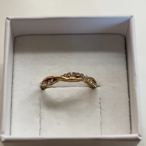 Guldfärgad ring med glittriga stenar - Snygg guldfärgad ring med en twistad design och glittriga stenar längs ovansidan. Perfekt för dig som gillar lite extra bling och vill sticka ut med en elegant men trendig accessoar.