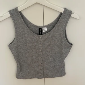 Grå ribbad linnetopp från H&M Divided - Grå croppad linnetopp från H&M Divided med ribbad struktur och snygga silverfärgade knappar framtill. Toppen är ärmlös och har en rundad halsringning, perfekt för en chill och stilren look. Lappen sitter kvar. 