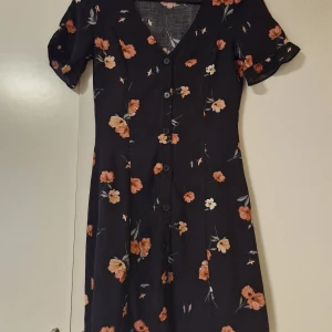 Svart blommig kortklänning med knappar - Supersöt svart kortklänning med rosa och vita blommor, v-ringning och knappar framtill. Klänningen har korta ärmar med liten volangkant och en lätt utställd passform. Perfekt för sommarens alla tillfällen!