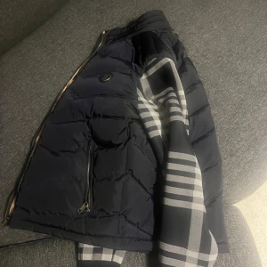 Mörkblå dunväst från Burberry M - Säljer en mörkblå dunväst från Burberry med klassiskt rutigt foder i beige, svart, vitt och rött. Västen har dragkedja framtill, två fickor med dragkedja och diskret Burberry-logga på ryggen. Perfekt för lager-på-lager och snygg detalj med rutigt mönster på insidan.