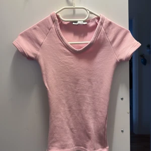 Rosa v-ringad topp från Zara - Söt ljusrosa topp från Zara med v-ringning och korta ärmar. Modellen är figurnära och har en enkel, stilren design utan mönster eller detaljer. Perfekt att matcha med jeans eller kjol för en fräsch look. 