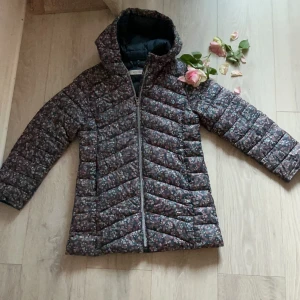 Blommig pufferjacka från MANGO 140 cm - Blommig pufferjacka från MANGO med dragkedja och huva. Jackan har mörk botten med små blommor i rött, vitt och grönt. Perfekt för kyliga dagar och har en skön, vadderad känsla. Långärmad och med snygg quiltad design.