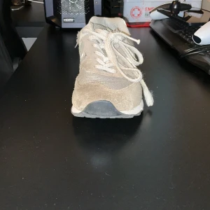 Grå New Balance sneakers i mocka - Snygga grå sneakers från New Balance med klassisk vit N-logga på sidan. Skorna har ovandel i mocka och mesh, snörning och platt sula. Perfekta för dig som gillar en clean och sportig stil. Passar till både jeans och shorts.