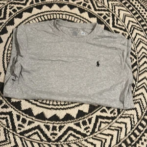 Grå t-shirt från Ralph Lauren - En stilren grå t-shirt från Ralph Lauren med klassisk rund hals och diskret broderad logga på bröstet. T-shirten är kortärmad och tillverkad i mjuk bomull, perfekt för en avslappnad look. Endast testad. Tveka inte på att höra ev dig vid minsta fundering.