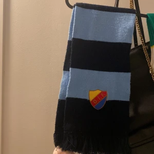 Randig Djurgården halsduk blå/svart - Säljer en klassisk halsduk från Djurgårdens IF med breda marinblå och ljusblå ränder. Halsduken har DIF:s klubbmärke broderat och fransar i ändarna. Perfekt accessoar för att visa ditt stöd på matchen!