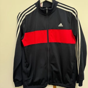 Adidas svart och röd vindjacka - Svart vindjacka från Adidas med en bred röd panel över bröstet och klassiska vita ränder längs ärmarna. Jackan har dragkedja framtill, ribbade muddar och Adidas-logga på bröstet. Perfekt för en sportig och avslappnad stil.