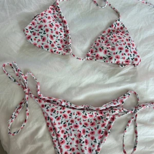 Blommig bikini med knytband - Aldrig använd! Supersöt bikini med vitt tyg och små rosa blommor över hela. Klassisk trekantsmodell på överdelen och knyt i sidorna på underdelen. Perfekt för stranden eller poolen. Tunt och smidigt material som torkar snabbt.