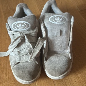 Beige Adidas Campus sneakers i mocka - Säljer ett par beige Adidas Campus sneakers i mocka med klassiska vita ränder och gummisula. Skorna har snörning och rund tå, samt Adidas logga på plös och häl. Perfekta för dig som gillar retrovibbar och streetstyle. helt okej skyck