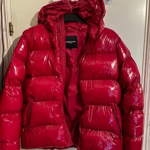 Röd pufferjacka från Lawrence Grey - Snygg röd pufferjacka från Lawrence Grey med glansig finish och huva. Jackan har dragkedja framtill och två fickor med dragkedja. Perfekt för kalla dagar när du vill sticka ut med en färgstark look.