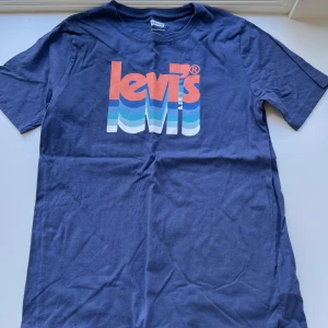Blå Levi's t-shirt med retrotryck - Snygg blå t-shirt från Levi's med stort retroinspirerat logotryck i orange, vitt och blått på bröstet. Klassisk passform, korta ärmar och rund hals. Tillverkad i mjuk bomull, perfekt för en chill och stilren look.