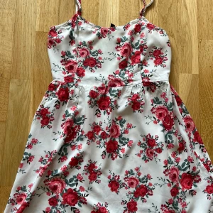 Vit blommig sommarklänning H&M Divided - Supersöt vit kortklänning från H&M Divided med röda och rosa blommor, smala axelband och smockad rygg. Funkar både som kortklänning eller längre top (beroende på hur lång man är). Lappen säger storlek 38 men jag upplever att den är liten i storleken och tror att den passar bättre på någon som normalt har s. Hör av dig vid frågor!😊