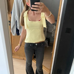 Ljusgul topp med breda axelband - Superfin ljusgul topp med breda axelband och rosettdetalj. Toppen har fyrkantig urringning och har fin passform. Helt ny, med prislappen kvar och såklart är oanvänd. 