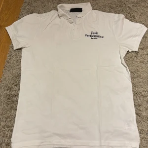 Vit pikétröja Peak Performance L - Klassisk vit pikétröja från Peak Performance i storlek L. Tröjan har korta ärmar, krage och knäppning framtill samt broderad logga på bröstet. Tillverkad i mjuk bomull som känns skön mot huden. Perfekt för en clean och sportig stil.