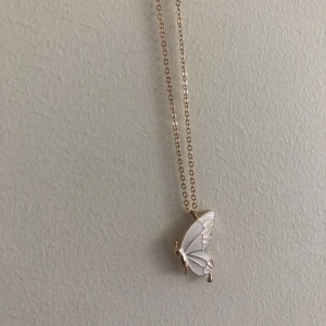 Guldhalsband med vit fjäril - Superfint halsband med tunn guldfärgad kedja och en söt vit fjäril som hänge. Fjärilen har detaljer i guld och ger ett elegant och lekfullt intryck. Perfekt accessoar för att lyfta vilken outfit som helst.