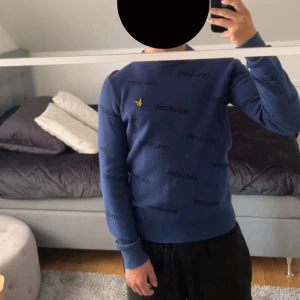 Blå stickad tröja från Lyle & Scott - Snygg blå stickad tröja från Lyle & Scott med svart logotyp-tryck över hela tröjan och den klassiska gula fågeln broderad på bröstet. Tröjan har rund halsringning, ribbade muddar och lång ärm. Perfekt för dig som gillar stilrena och trendiga plagg.