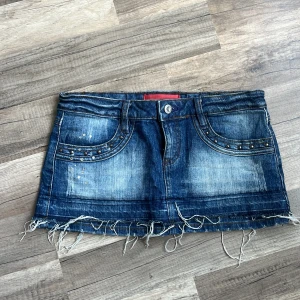 Blå mini jeanskjol med pärlor - Säljer en blå jeanskjol i kort modell med ”sliten style” . Kjolen har coola nitar och paljettdetaljer både fram och bak, samt klassiska fickor. Perfekt för dig som gillar en edgy och trendig stil.