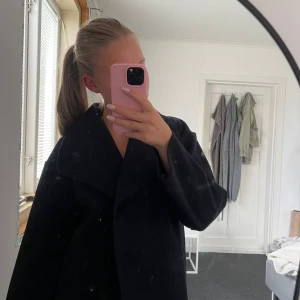 Svart kappa från Gina Tricot - Stilren mörkblå kappa från Gina Tricot med bred krage och knappar framtill. Jackan har en rak passform och är i ett mjukt material som känns lite ulligt. Perfekt för dig som gillar en klassisk look med modern twist.