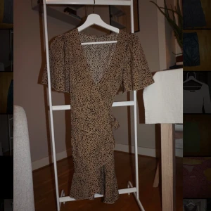 Beige omlottklänning med prickar - Supersöt kort omlottklänning i beige med svarta prickar. Klänningen har v-ringning, volangkant nertill och vida korta ärmar. Knyt i midjan för en snygg passform. Perfekt för sommarens alla tillfällen!