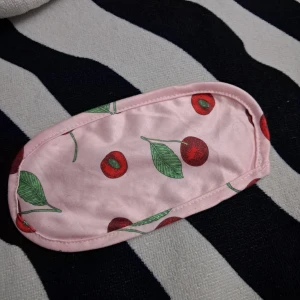 Rosa sovmask med körsbärsmönster - Söt rosa sovmask med körsbär och gröna blad som print. Masken har en mjuk känsla och elastiskt band på baksidan för att sitta bekvämt. Perfekt för att blocka ljus när du vill vila eller resa. - fint skick- använd endast en gång 