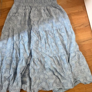 Ljusblå långkjol med mönster - Superfin ljusblå långkjol med vitt mönster och smock i midjan. Kjolen har volanger och ett luftigt fall, perfekt för varma dagar. Snygg att styla med en enkel topp och sneakers.