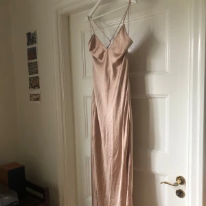 Champagnefärgad långklänning med tunna band - En elegant långklänning i champagnefärg med tunna axelband som korsas i ryggen. Klänningen har v-ringning och är tillverkad i ett glansigt, slätt material🩷