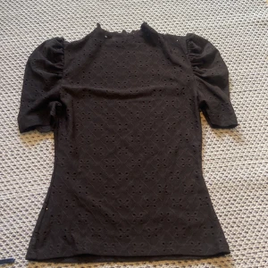 Svart blus med puffärm  - Supersnygg svart blus med puffiga ärmar och broderade hålmönster över hela tyget. Lite högre halsringning och figurnära passform ger en stilren look. Perfekt att styla med jeans eller kjol för en trendig outfit.