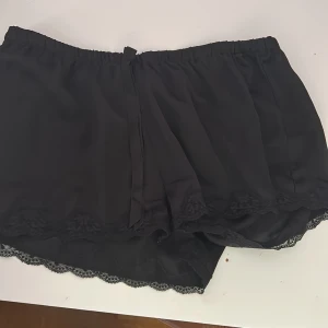 Svarta satin spetskantade pyjamas shorts - Svarta shorts från H&M med elastisk midja och snygg spetskant nertill. Lätta och flowiga i materialet, perfekta för varma dagar eller som mysplagg. Klassisk look med en feminin touch tack vare spetsdetaljerna.