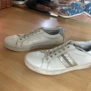 Vita sneakers med guldiga detaljer - Snygga vita sneakers med guldiga och silvriga detaljer på sidan och guldiga öljetter. Skorna har snörning och en platt sula. Materialet är troligtvis syntet med inslag av skinnimitation. Perfekta för dig som gillar stilrena men ändå unika sneakers.