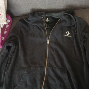 Svart hoodie från Converse med dragkedja - Svart hoodie från Converse med klassisk logga på bröstet. Tröjan har huva med snörning, hel dragkedja framtill och två stora fickor. Perfekt för en chill och sportig stil. Tillverkad i mjuk bomullsblandning för extra komfort.