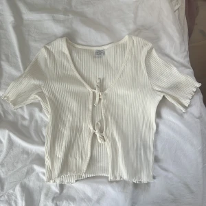 Vit ribbad knyttopp från Lindex - Supersöt vit ribbad blus från Lindex med korta ärmar och två knyt framtill. Toppen har dock en liten fläck  på framsidan men säkert går bort i tvätten!   Materialet är mjukt och stretchigt, vilket gör den extra bekväm.